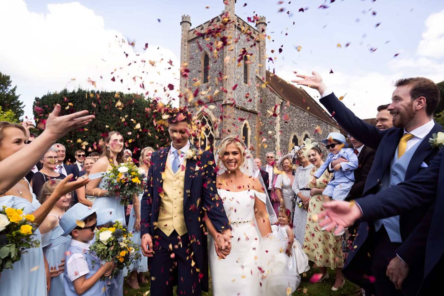 Joyful newlyweds walk through colorful confetti shower outside historic stone church surrounded by cheering wedding guests.
