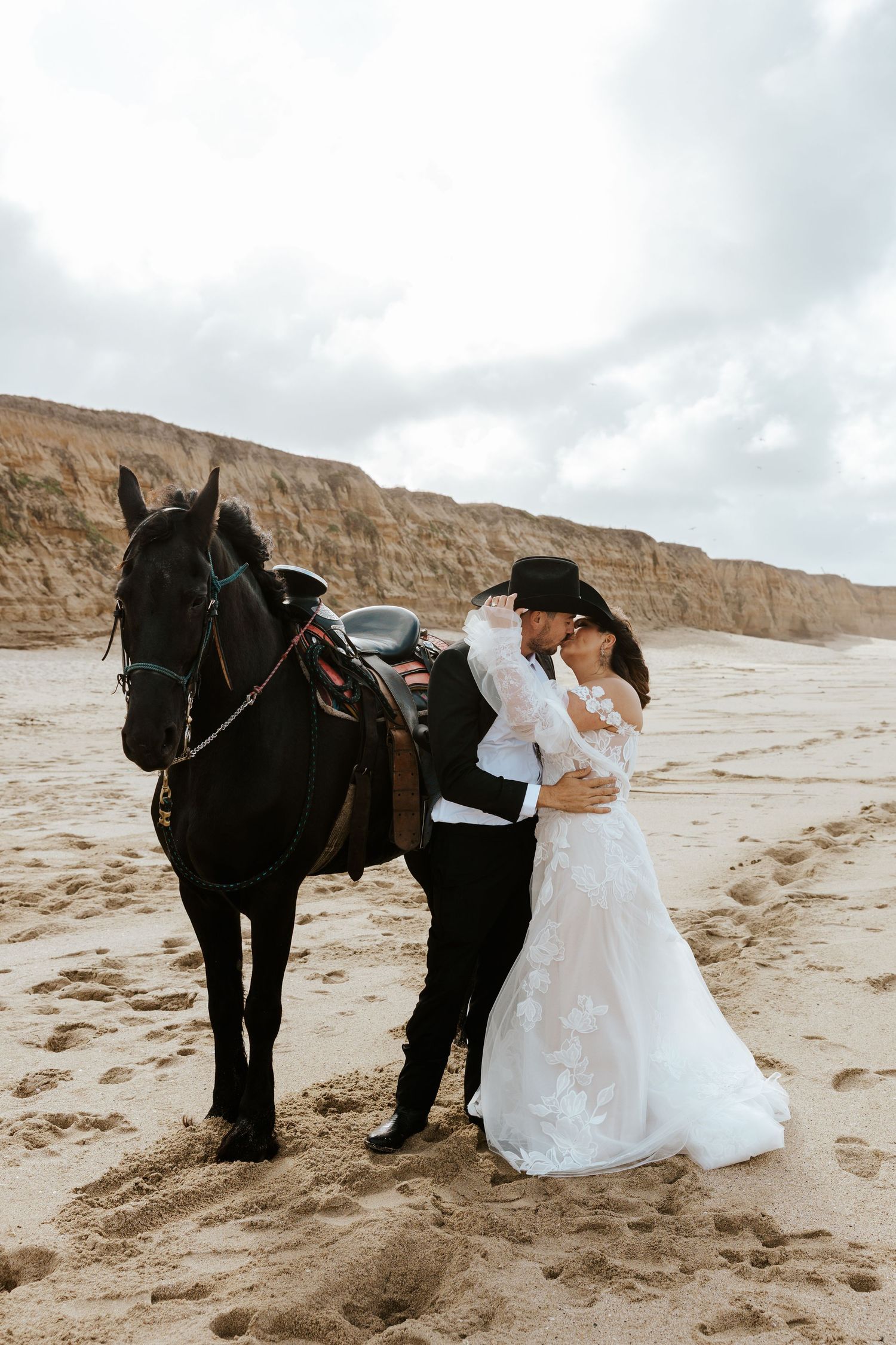 romantic candid couple kissing while standing next to a black horse on the beach in Half Moon Bay during Bay Area elopement