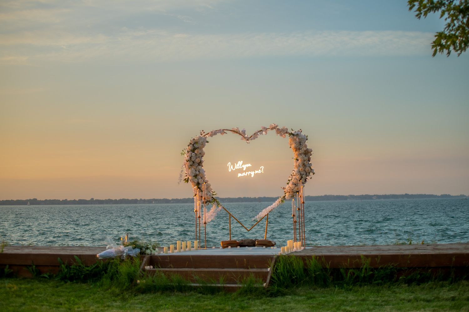 Heart-shaped floral wedding arch with neon sign by the waterfront at sunset.