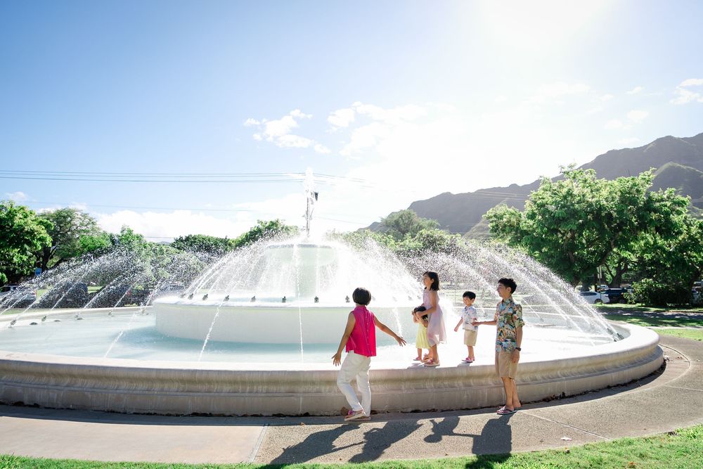 Family enjoying a large outdoor fountain in a sunny park with green trees and mountains in the background.