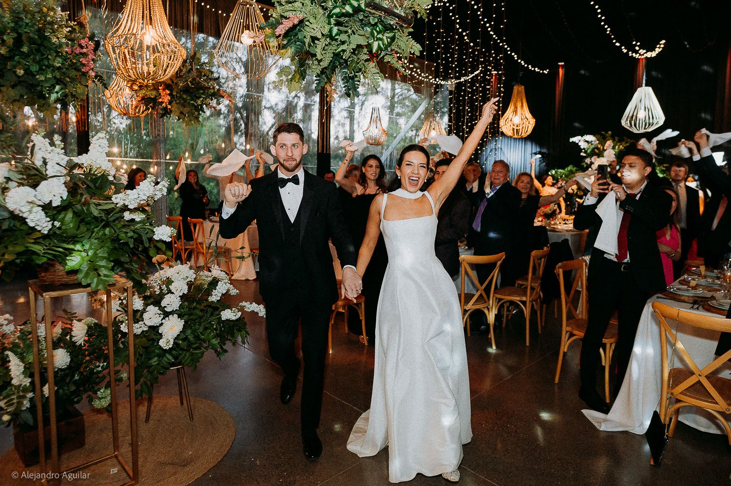 Bride and groom make grand entrance at elegant wedding reception with lush greenery, string lights, and cheering guests.