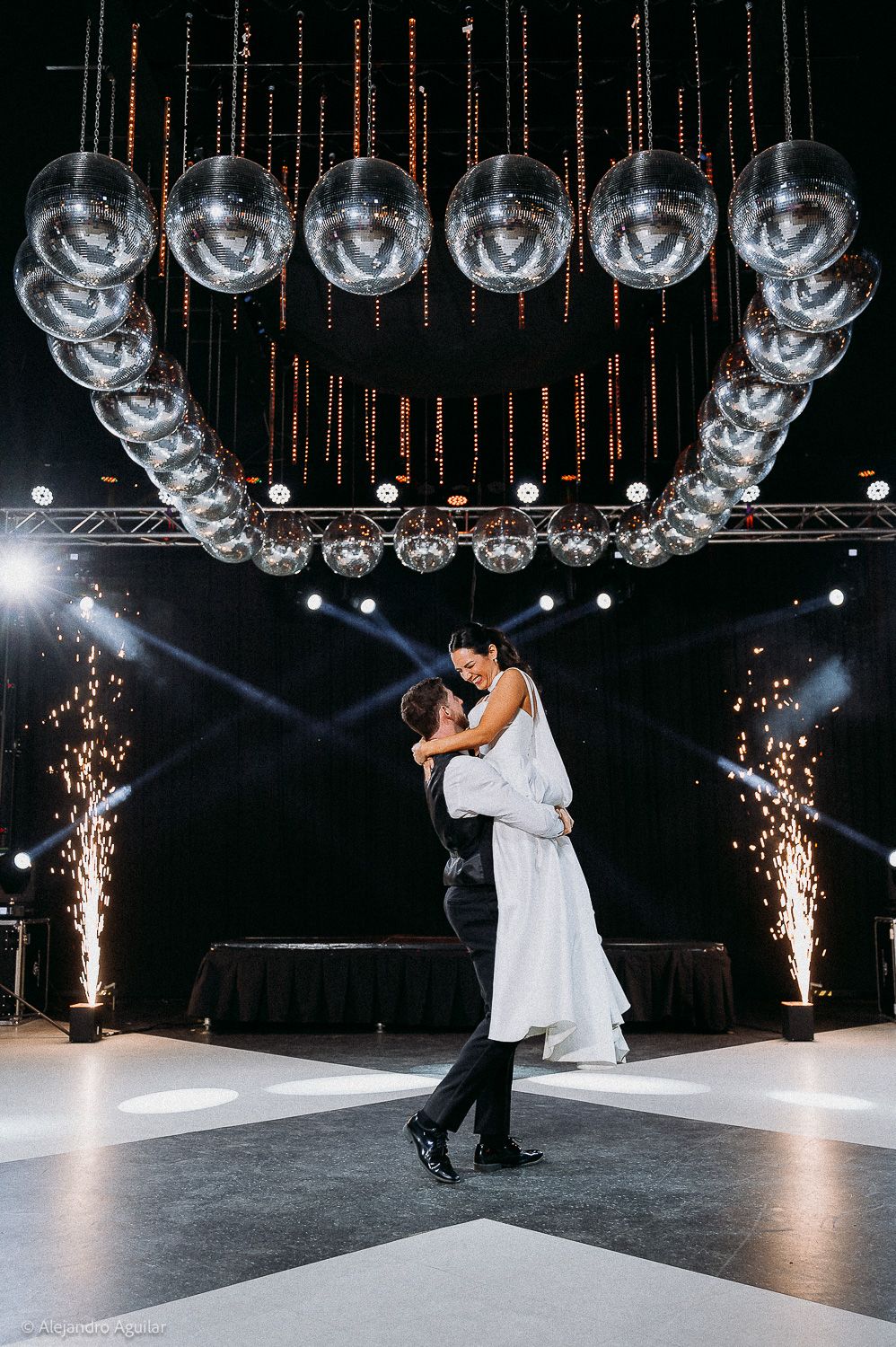 Groom lifts bride on dance floor under disco ball chandelier with sparkling fireworks at elegant wedding reception.