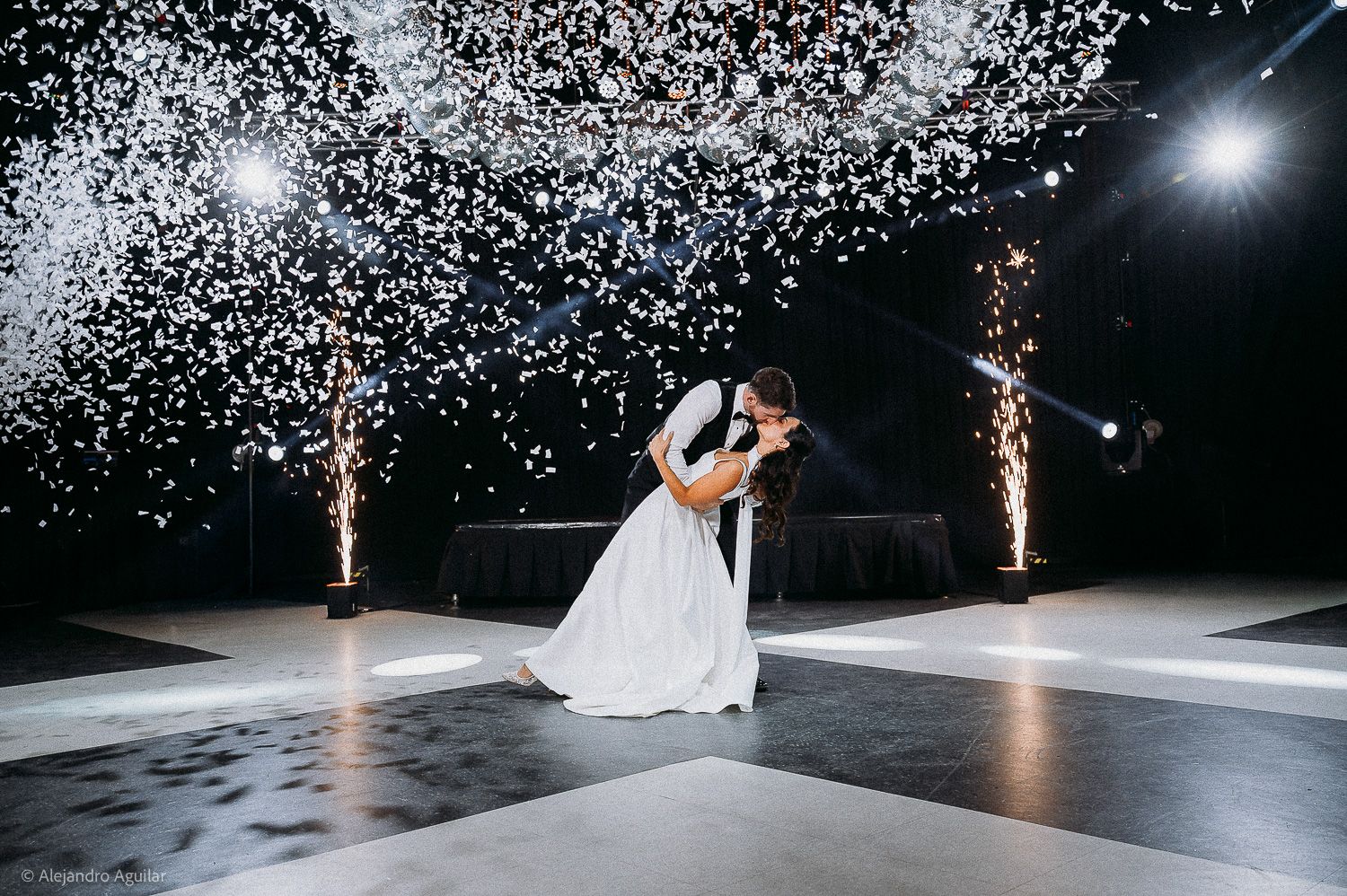 Bride and groom share romantic first dance under cascading white confetti with sparkling fountains at elegant wedding reception.