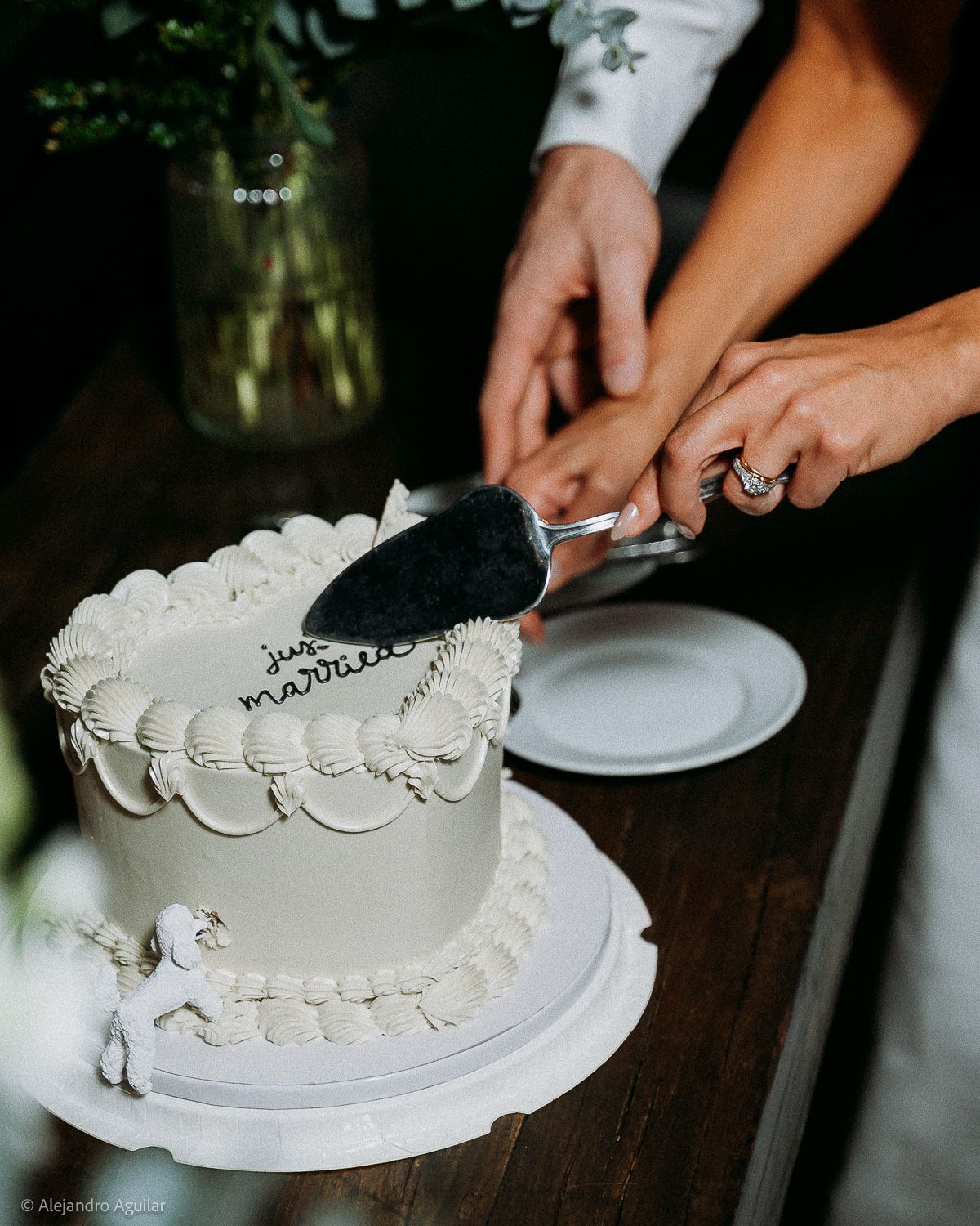 Couple cutting a white 'Just Married' wedding cake with a silver server on a white cake stand.
