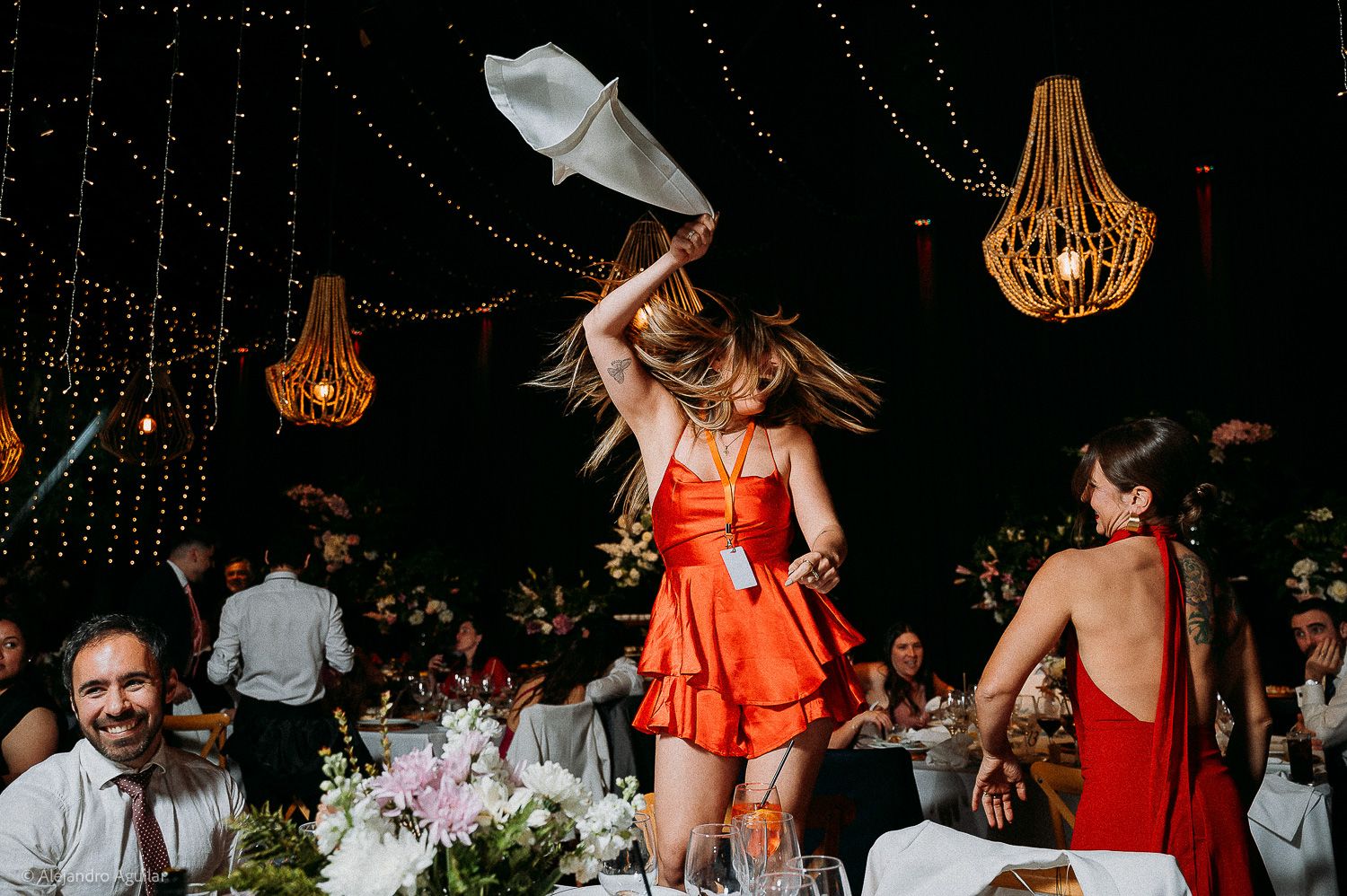 Woman in orange dress dancing joyfully at elegant wedding reception with chandeliers and floral centerpieces.