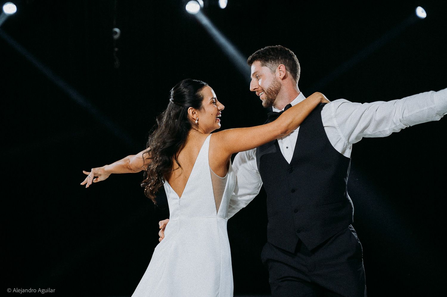 Bride and groom share a joyful first dance, arms outstretched, smiling at each other under dramatic stage lighting.