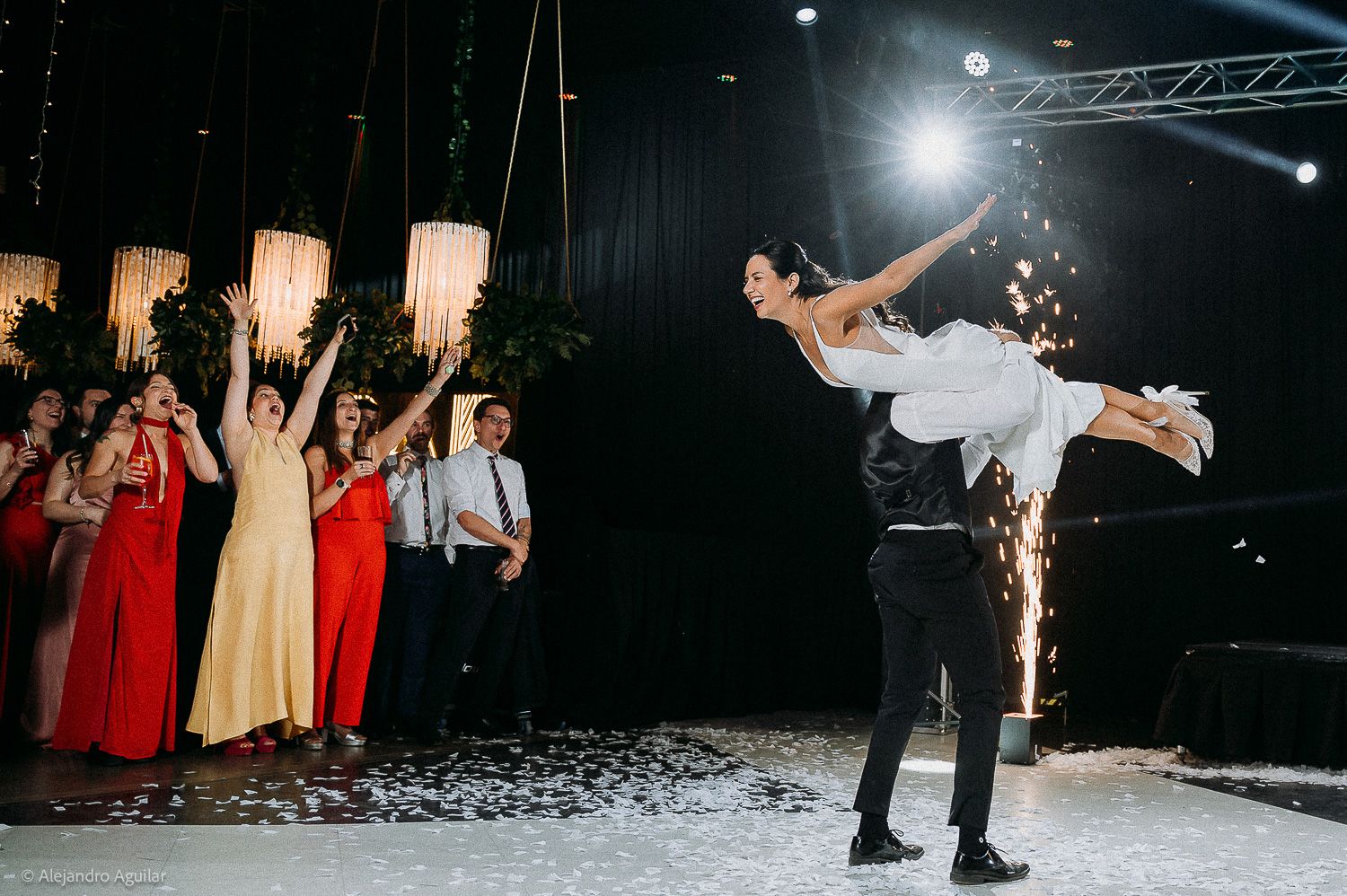 Bride lifted by groom during first dance at wedding reception with guests in red dresses cheering with sparklers.