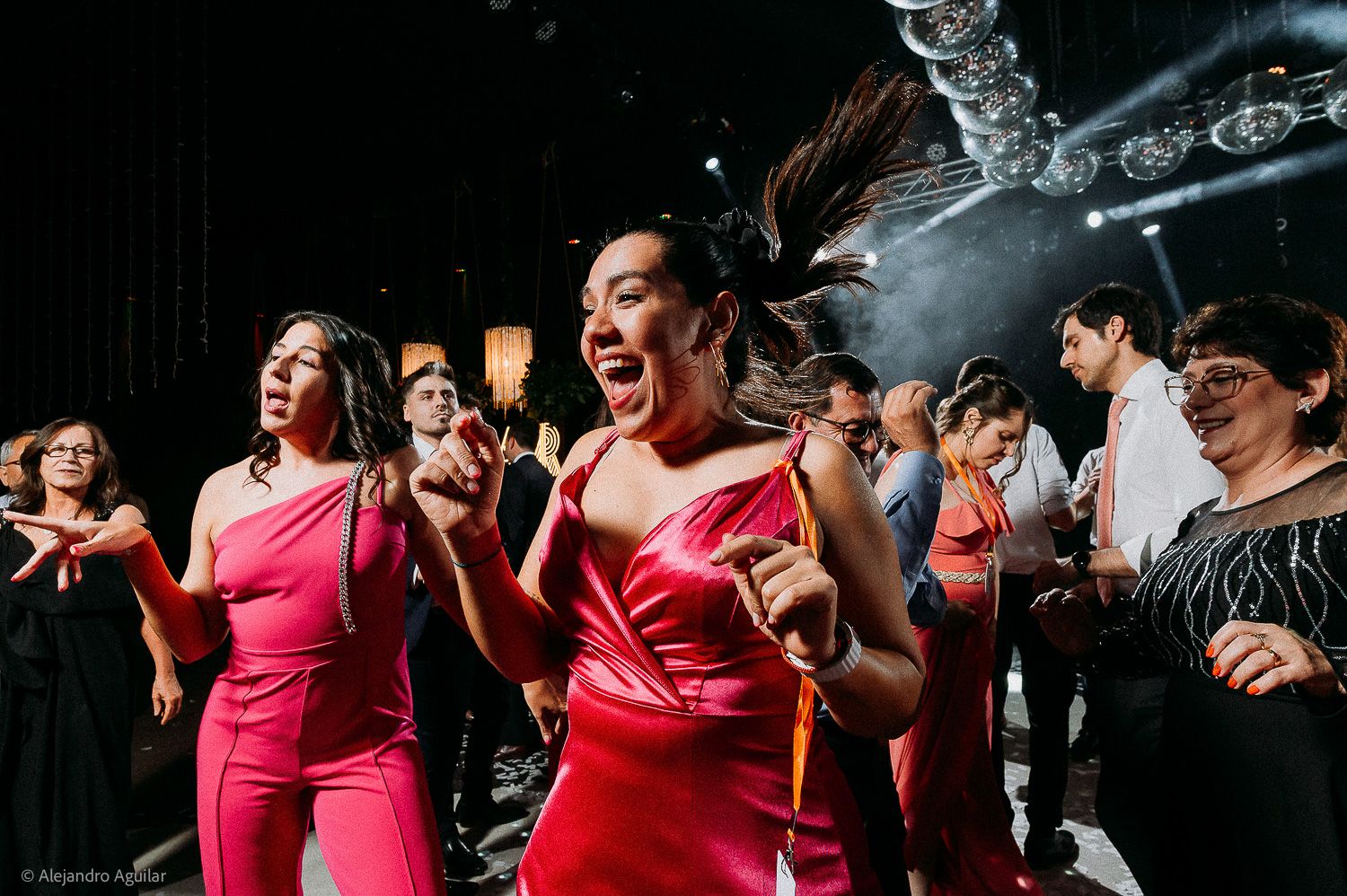 Women in red dresses dance energetically at a lively party event with stage lighting.