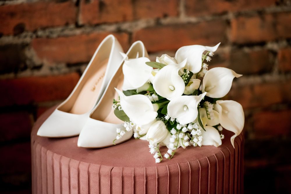 White bridal heels and calla lily bouquet with baby's breath displayed on a pink ribbed surface against brick wall.