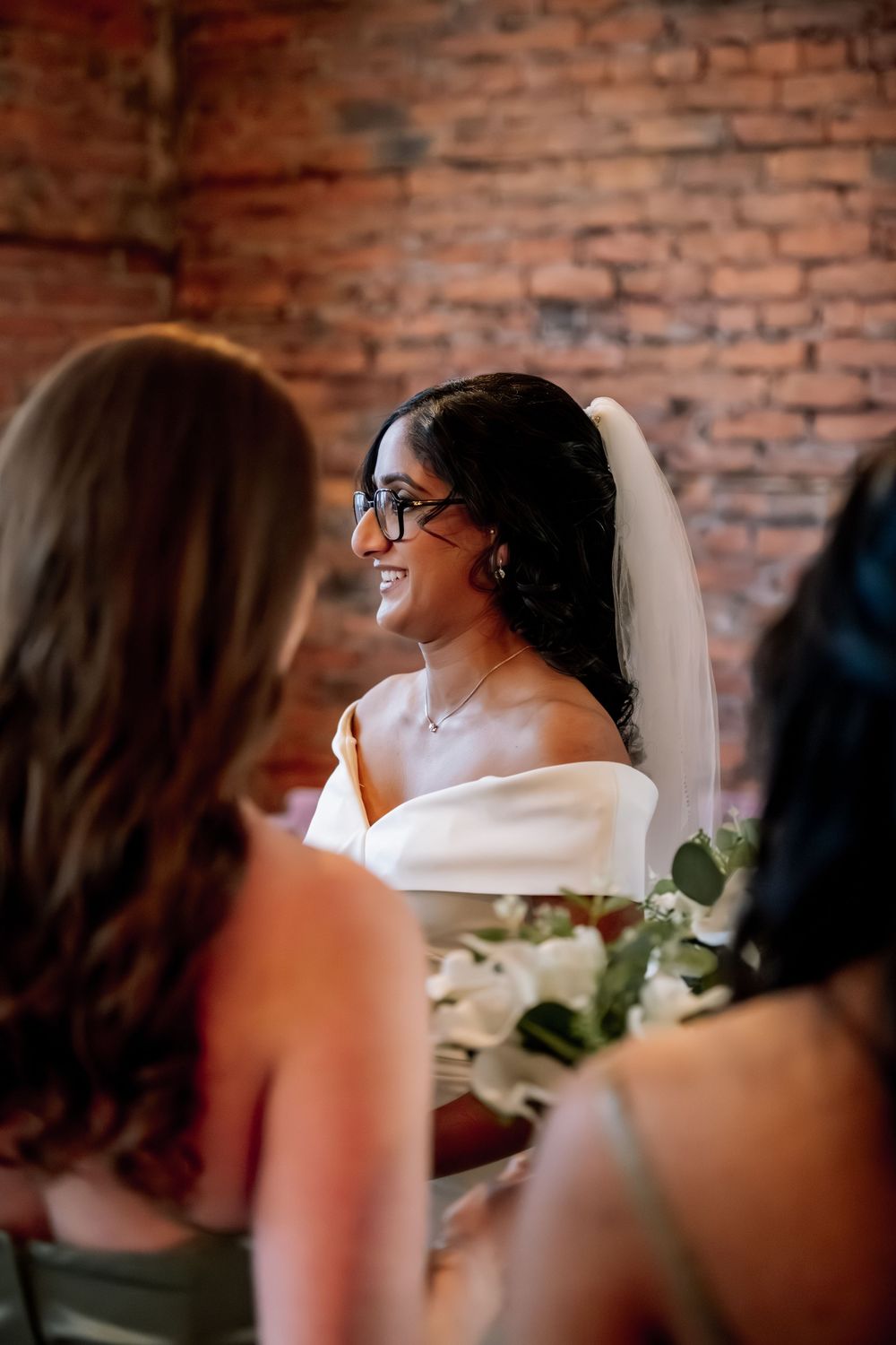 Smiling bride wearing glasses and veil holds white bouquet at indoor wedding ceremony with brick wall backdrop.