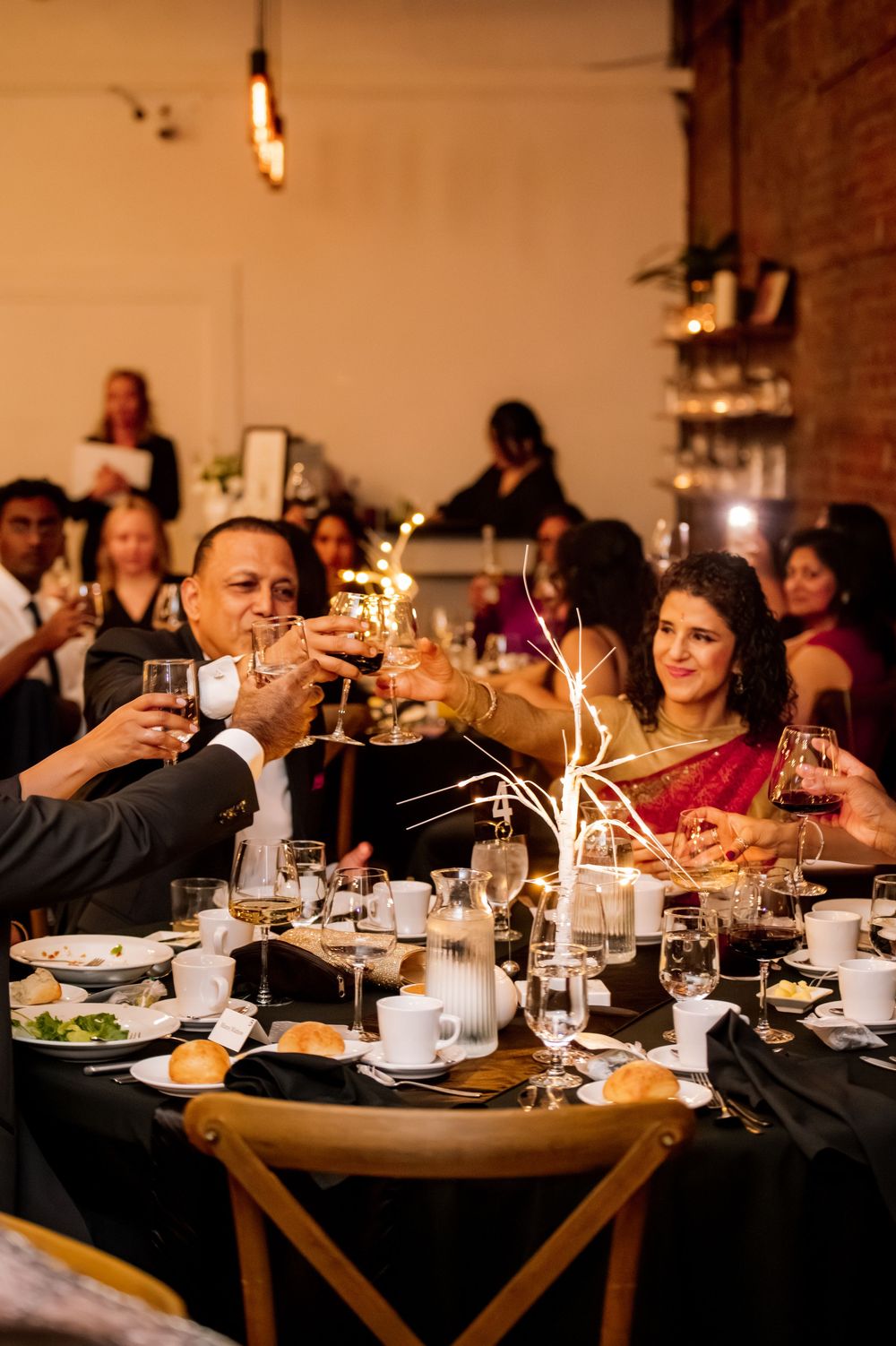 Wedding guests raise champagne glasses in a toast at elegant reception dinner with warm ambient lighting.