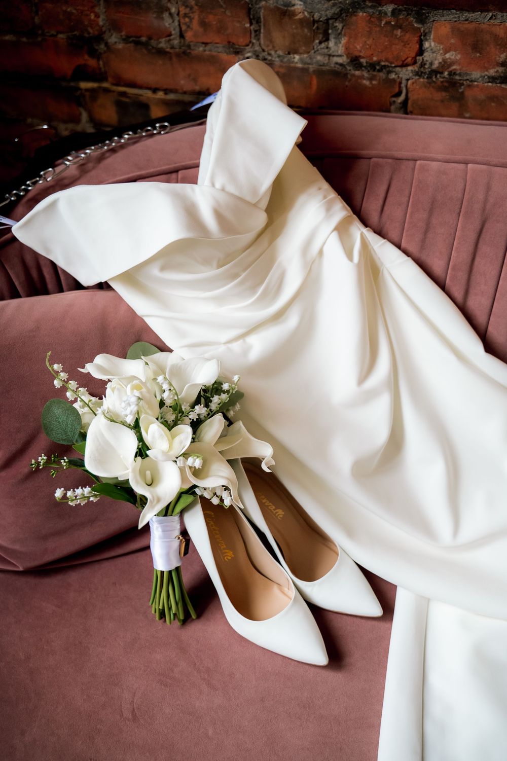 Elegant bridal flat lay featuring a white satin wedding dress, white heels, and a calla lily bouquet on a pink velvet sofa.