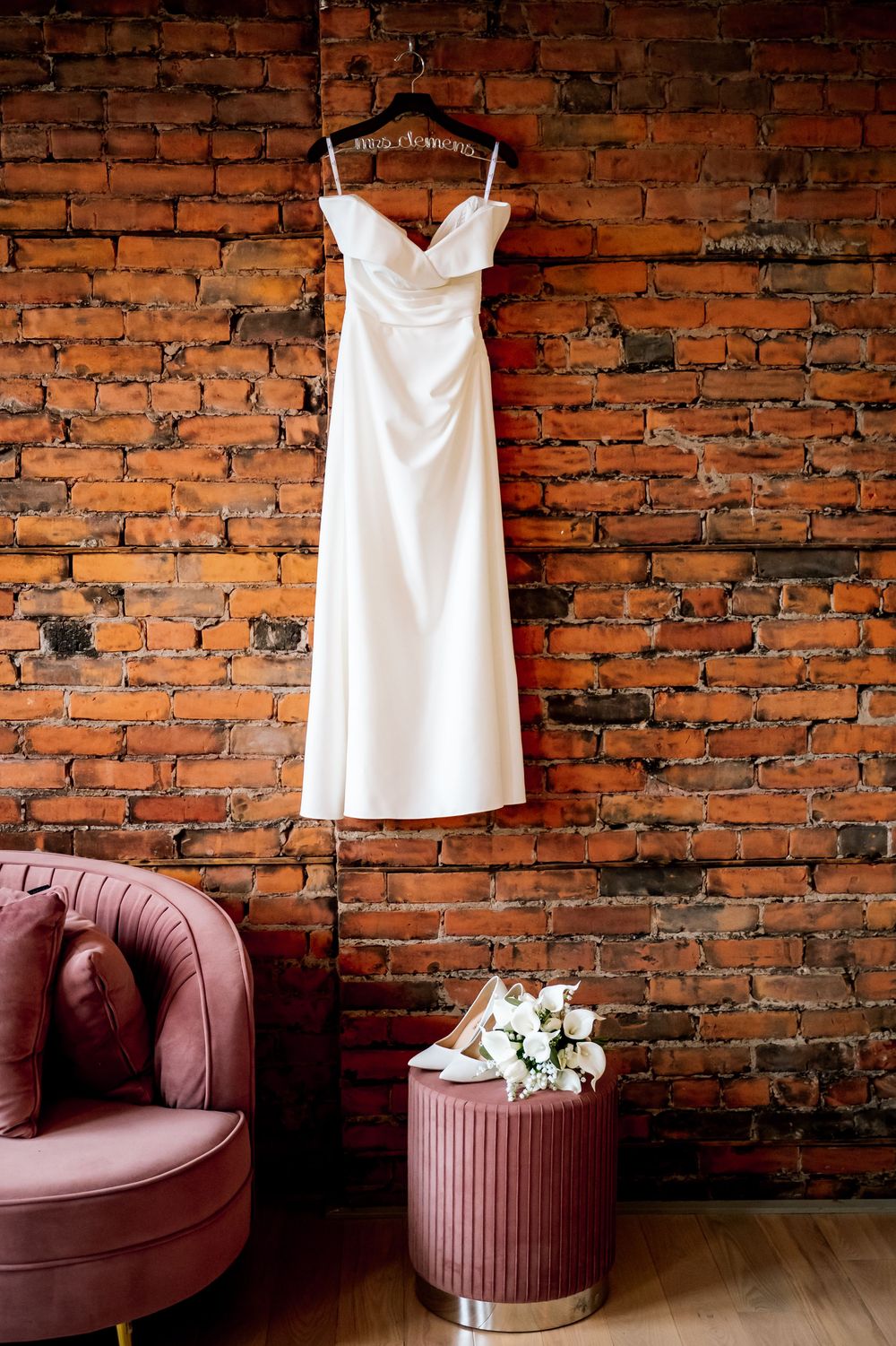 White satin wedding dress hanging on brick wall above bridal heels and bouquet beside pink velvet chair.