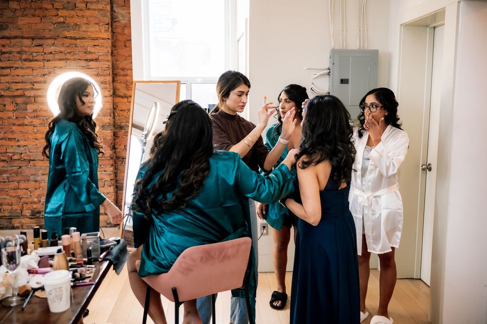Bridesmaids in robes getting makeup done in a bright studio with brick walls and vanity mirror.
