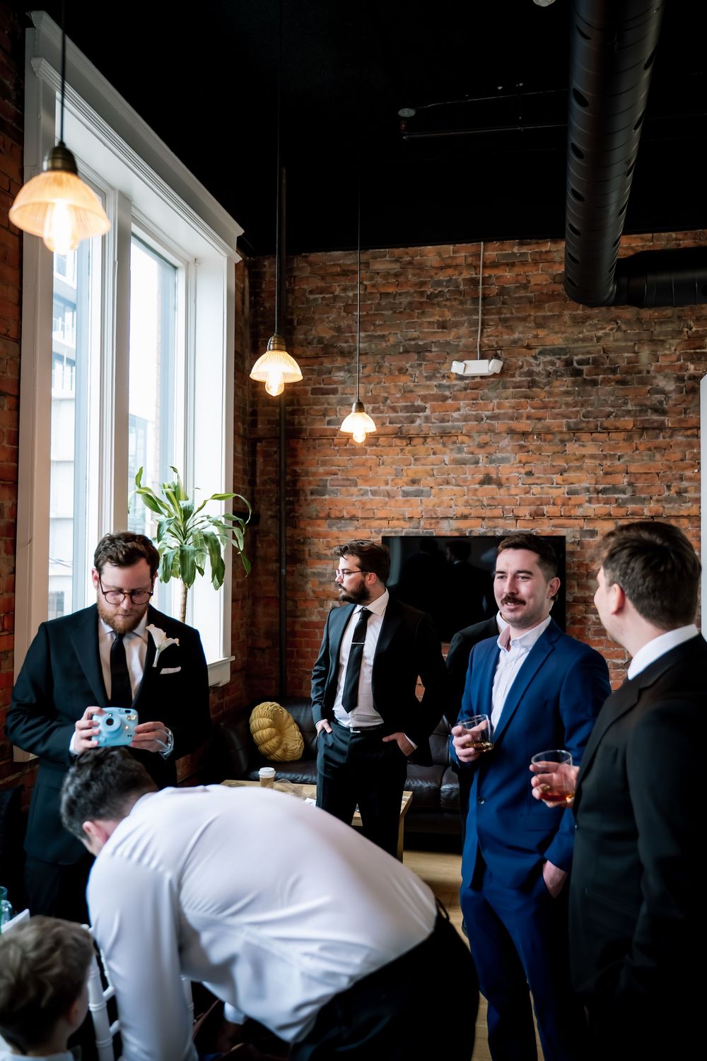 Groomsmen in suits gather in a rustic brick-walled venue with pendant lights before a wedding ceremony.