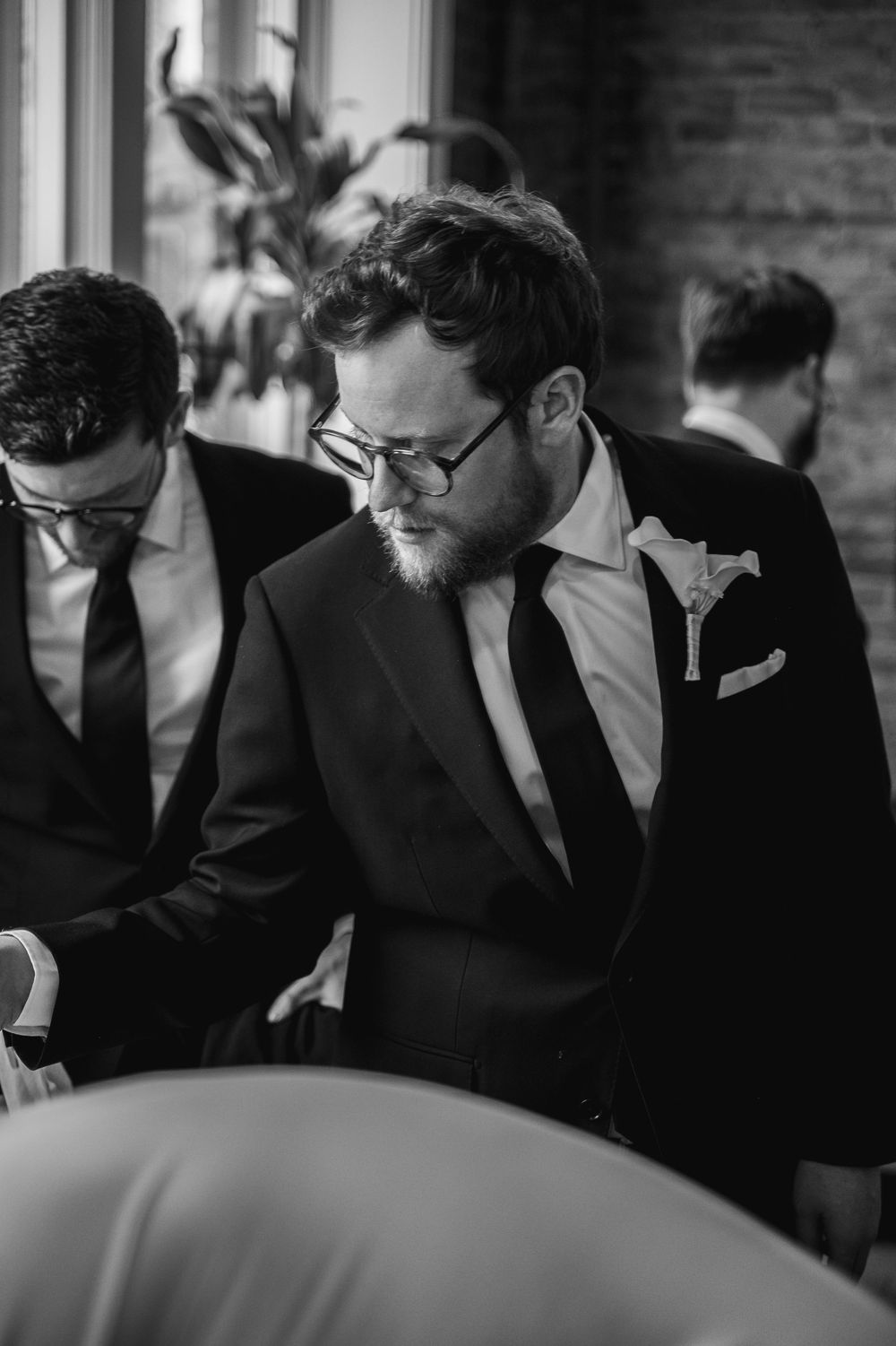 Groom in black tuxedo with boutonniere and round glasses looks down during wedding preparations.