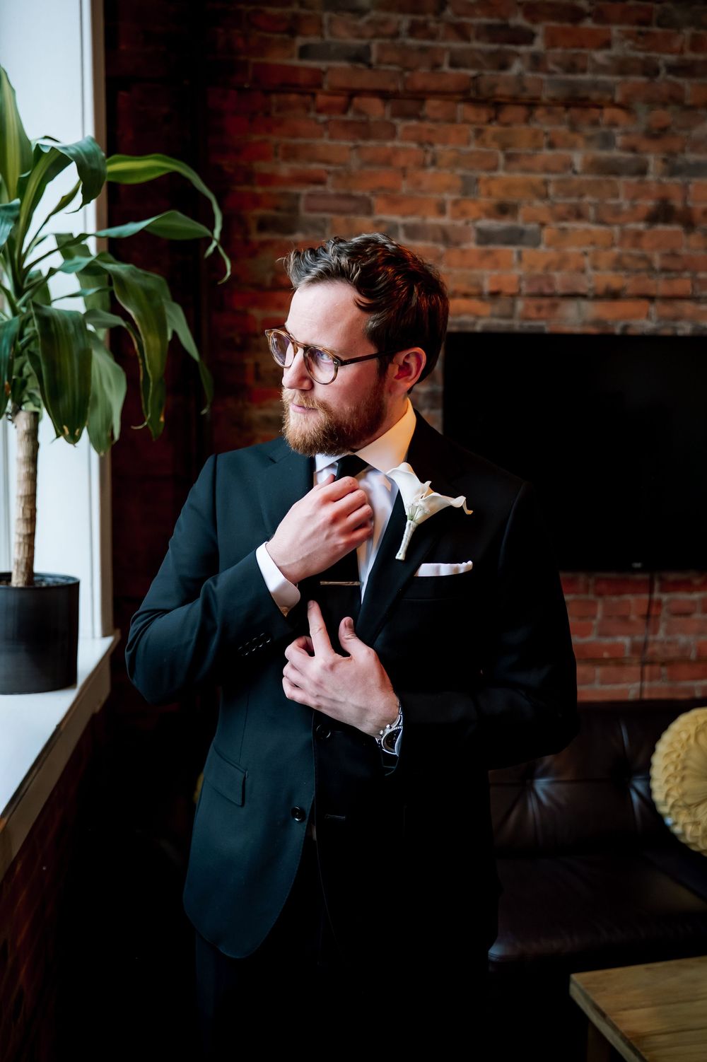 Bearded groom in dark suit adjusting tie with boutonniere near brick wall and green plant.