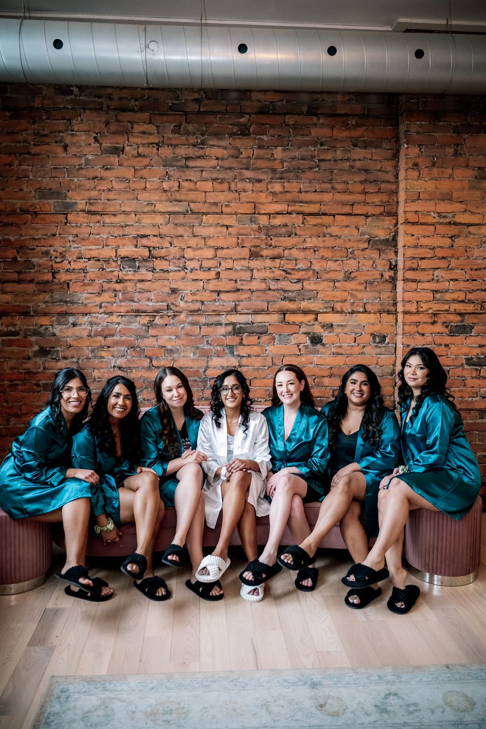 Bride in white robe with bridesmaids in teal satin robes and black slippers sitting against brick wall.