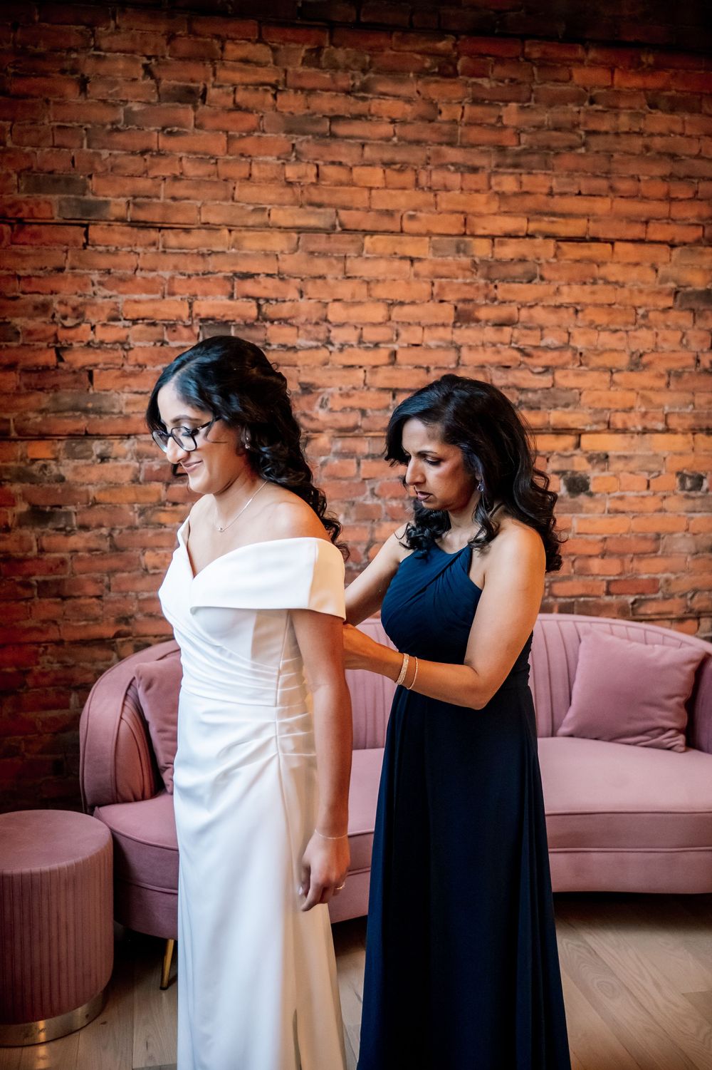 A bridesmaid in navy blue helps a bride in a white off-shoulder gown prepare in a brick-walled venue.