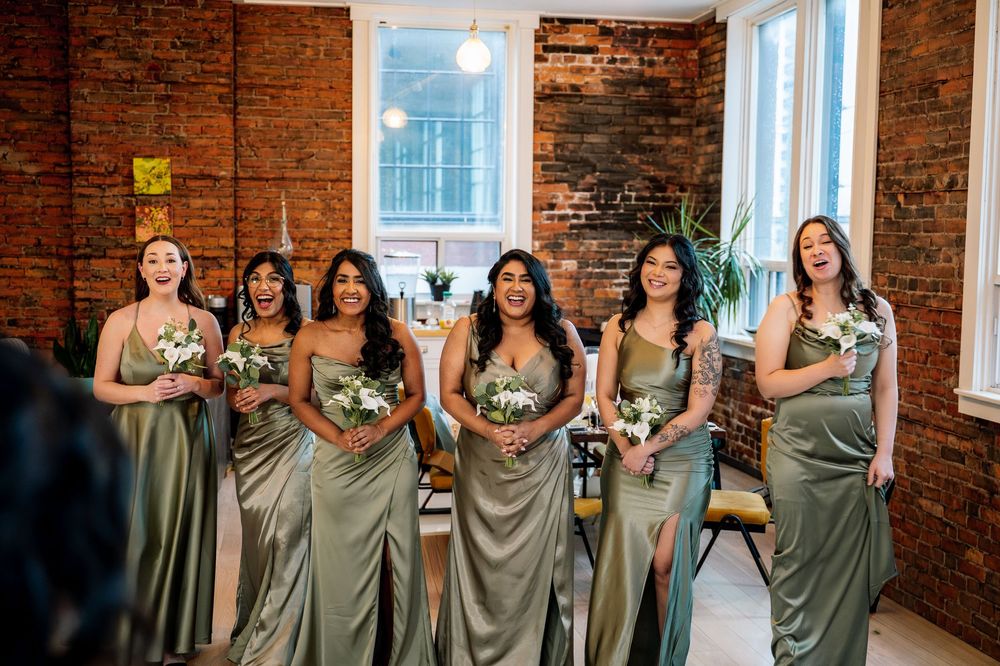 Five bridesmaids in sage green satin gowns holding white bouquets laugh together in a brick-walled wedding venue.