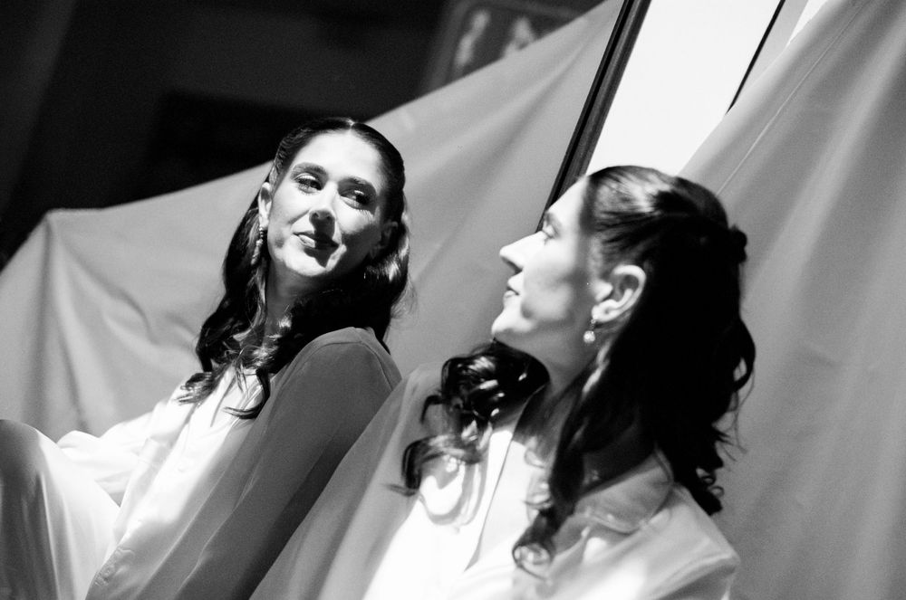 Two women in white dresses look upward dramatically in a black and white theatrical performance photo.