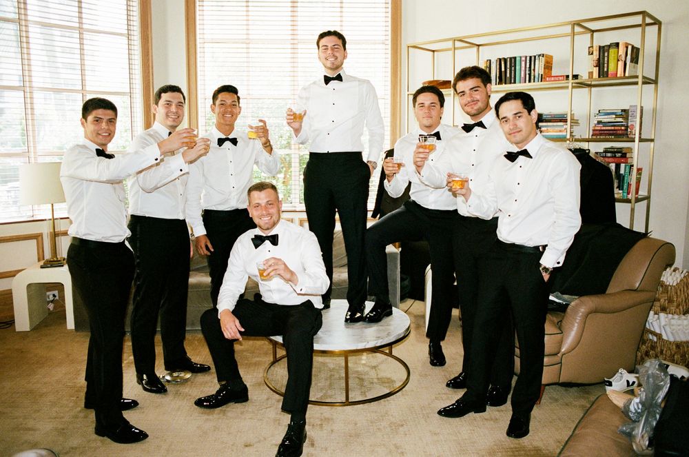 Groomsmen in black tuxedos and bow ties raising glasses for a toast in an elegant indoor setting.
