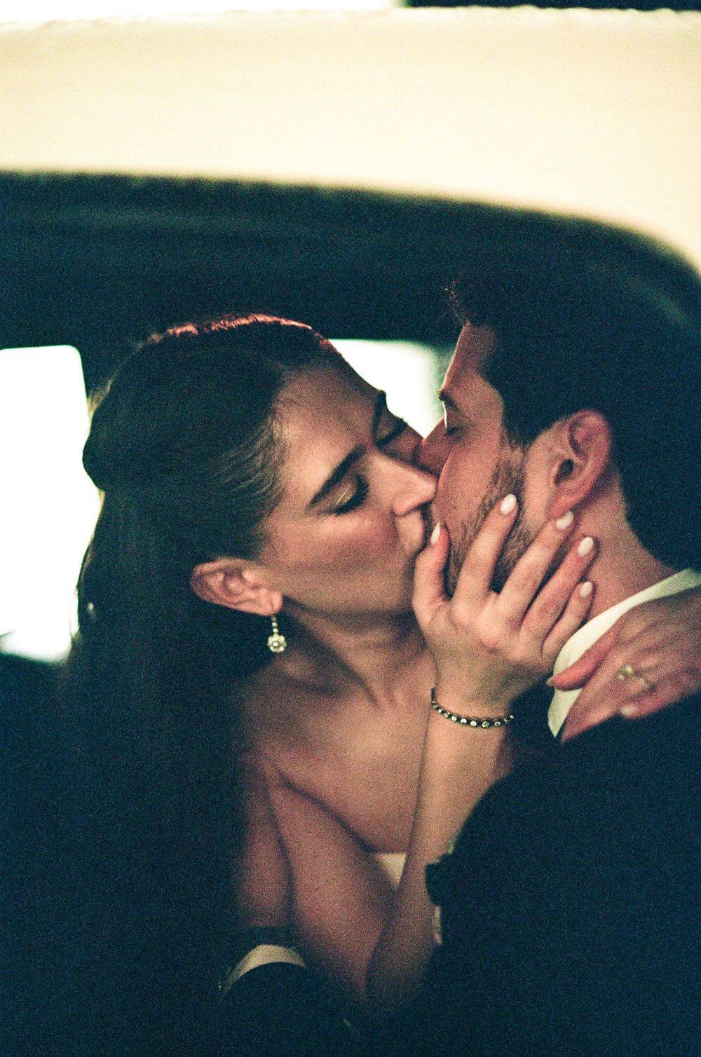 Couple sharing a romantic kiss inside a car, woman wearing elegant earrings and bracelet in a wedding setting.