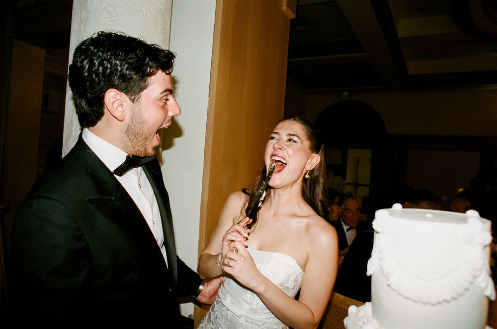 Bride and groom laughing while cutting white wedding cake, groom in tuxedo, bride in strapless gown.