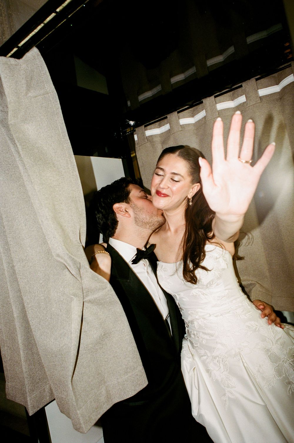 A bride and groom share a romantic kiss in a photo booth, with the bride raising her hand to show her wedding ring.