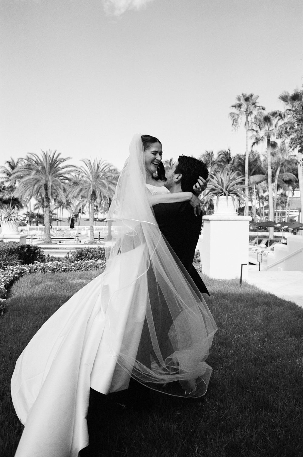 Black and white photo of a groom lifting his smiling bride in a wedding gown and veil outdoors among palm trees.