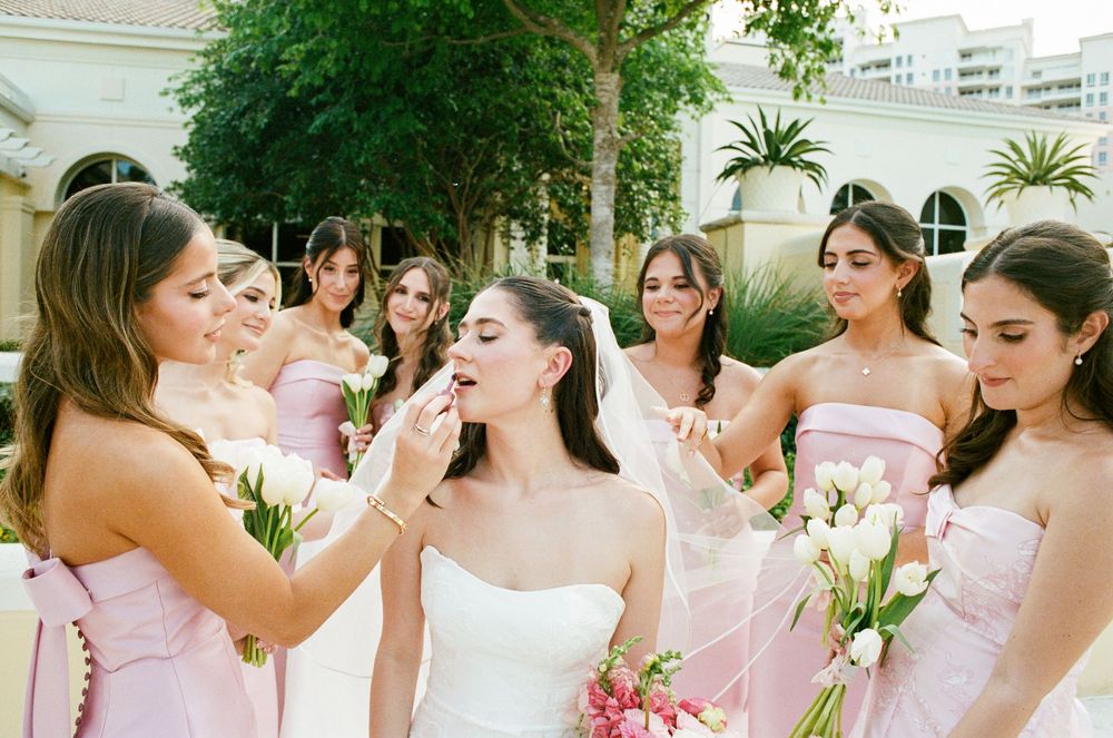 Bride in white gown surrounded by bridesmaids in pink dresses holding white calla lily bouquets outdoors.