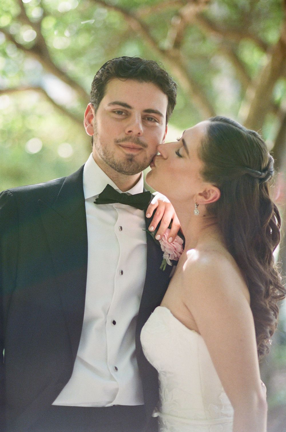 Bride kissing groom on cheek in wedding attire, tuxedo and white gown, outdoors under green trees.