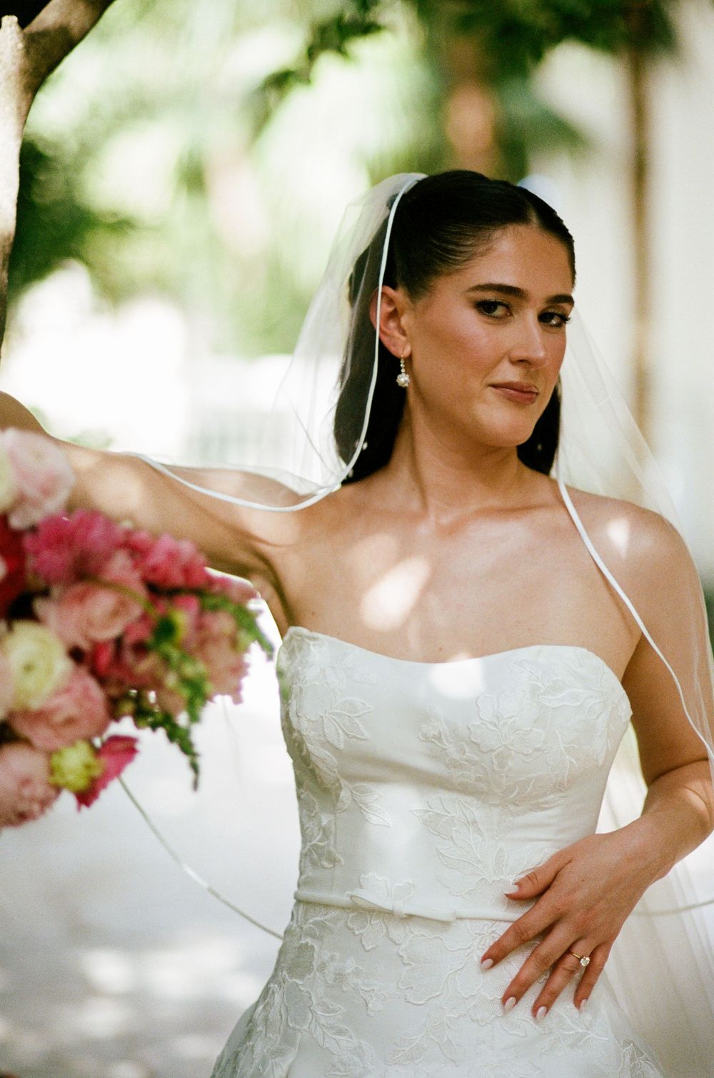 Elegant bride in strapless lace wedding gown and veil holds pink bouquet outdoors in soft natural light.