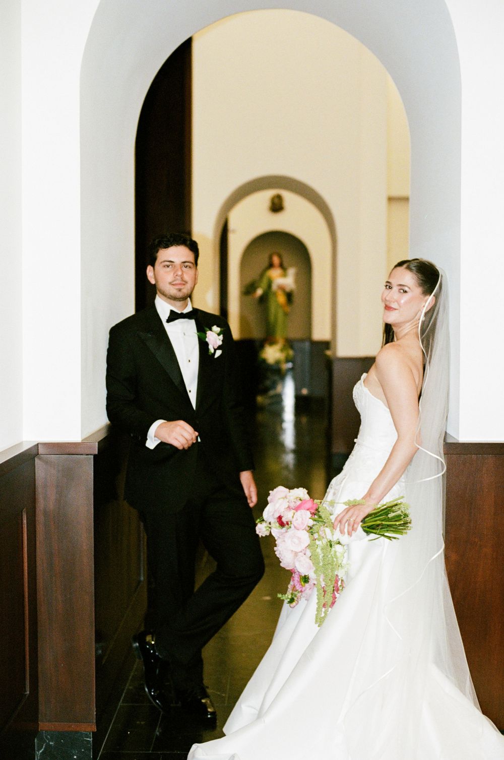 Bride and groom pose in elegant church hallway, she holds pink bouquet in white gown, he wears classic black tuxedo.