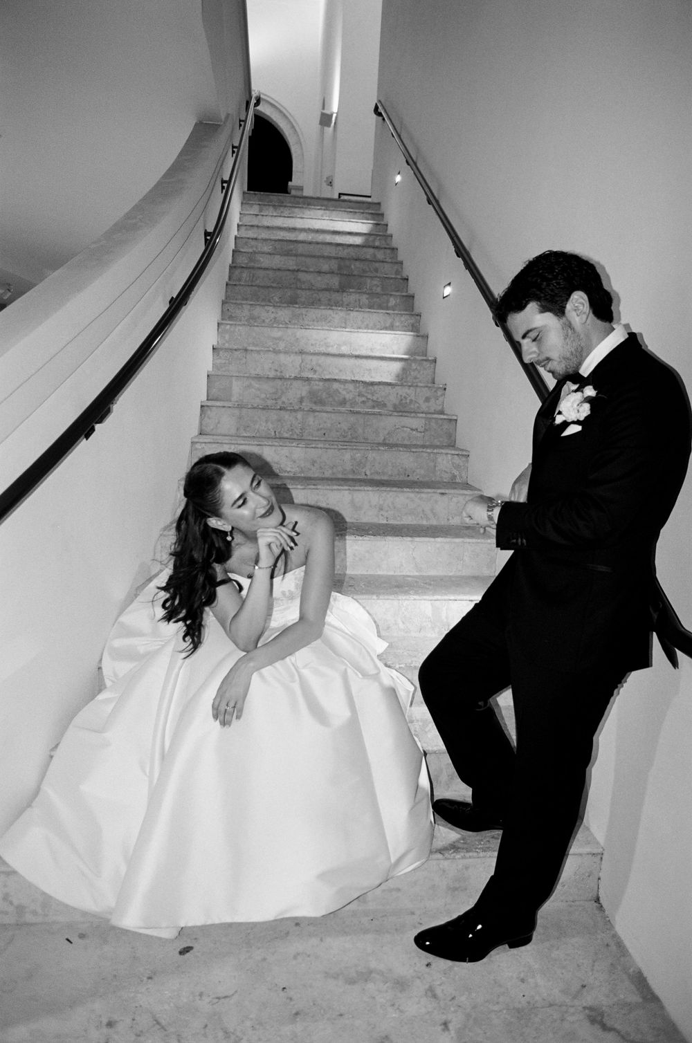 Black and white wedding photo of bride in gown and groom in tuxedo sitting on elegant staircase steps.