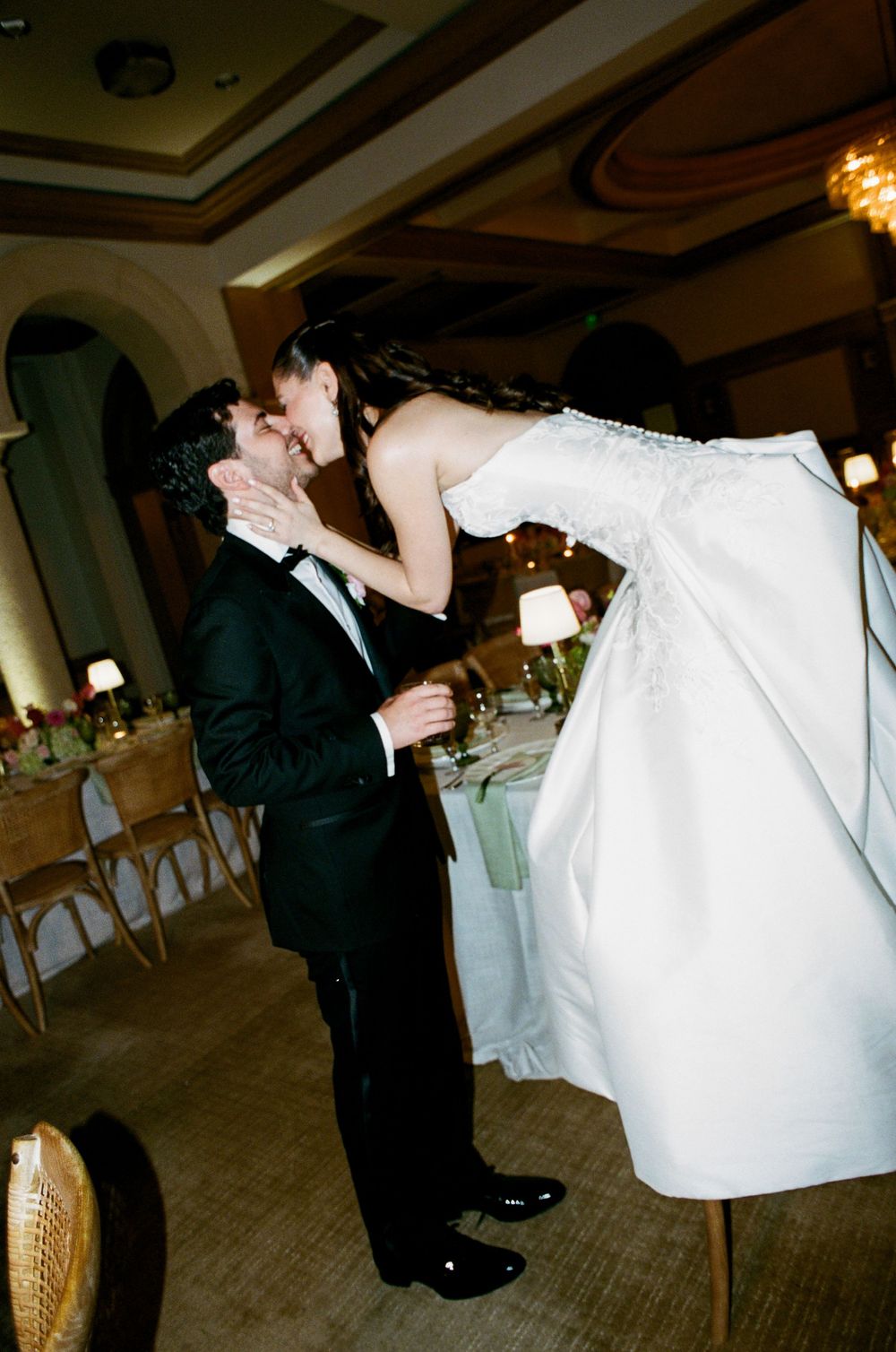 Bride and groom share a romantic kiss at their wedding reception, with the groom lifting his bride in a white gown.