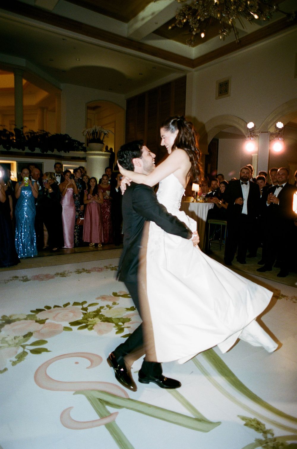 Groom lifts bride in white gown during first dance at elegant wedding reception with guests watching.
