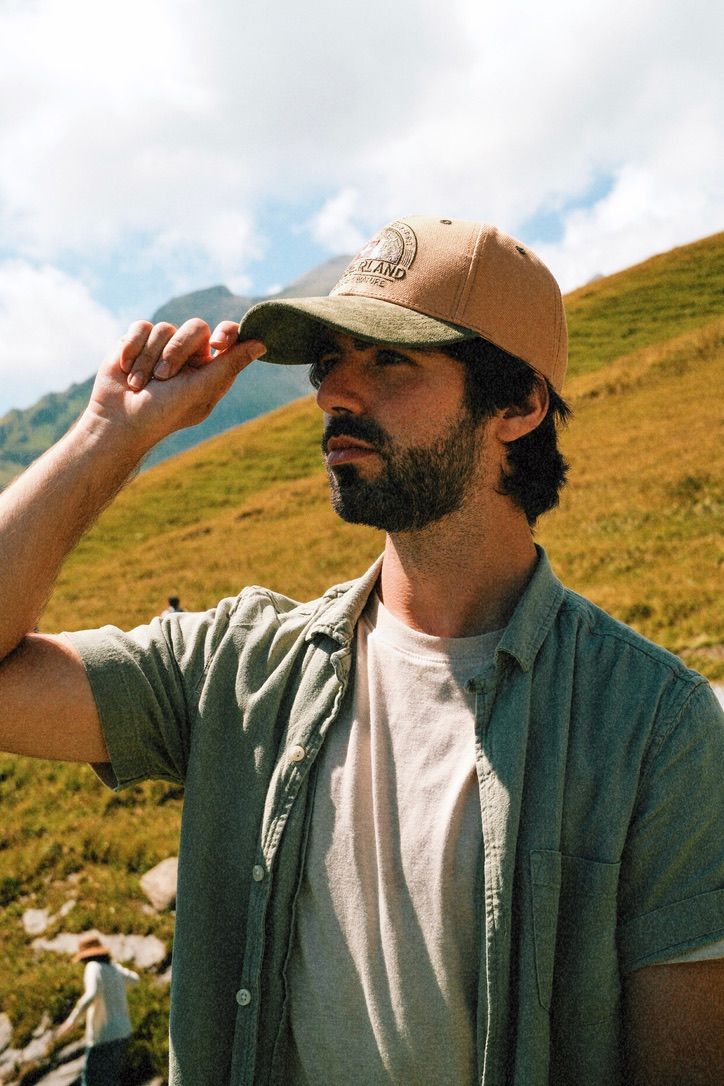 Bearded man adjusting tan baseball cap on green mountain hillside on cloudy day.