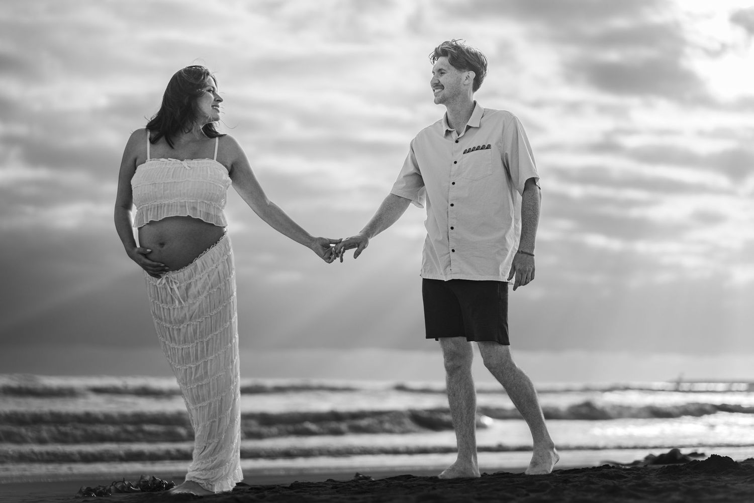 Pregnant couple holding hands on a beach at sunset, woman in white dress showing baby bump, man in white shirt. Oceanside, CA
