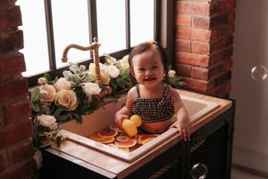 A smiling baby girl sits in a vintage sink surrounded by white flowers and citrus fruit slices in a charming photo shoot.