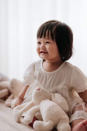 Smiling Asian toddler girl in white dress holding stuffed bunny toy, sitting on bed with soft light background.