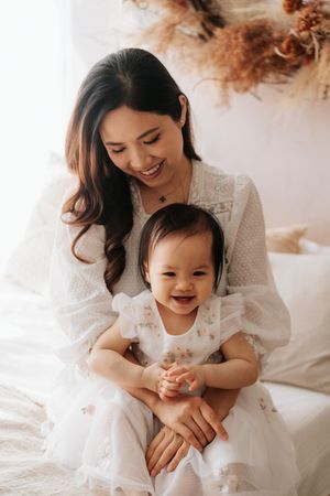 A smiling Asian mother in a white dress holds her laughing baby girl, both dressed in white, in a bright, cozy room.