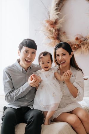 Happy Asian family portrait with parents and baby girl in white dress, smiling together in a bright studio setting.