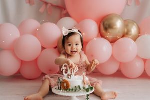 Baby girl in pink tutu smashing her first birthday cake surrounded by pink and gold balloons.