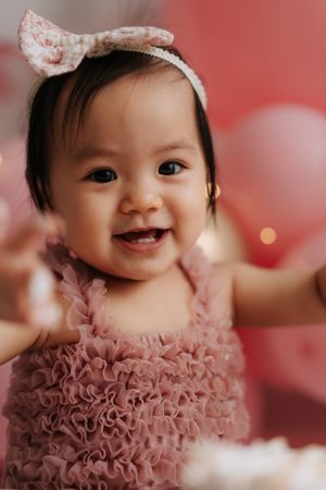 Smiling baby girl in pink ruffled dress with floral bow headband at first birthday party with pink balloons.