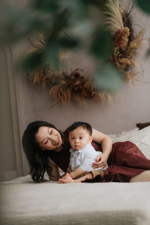 A mother smiles warmly while lying on a bed with her baby boy, surrounded by dried floral decor in a cozy room.