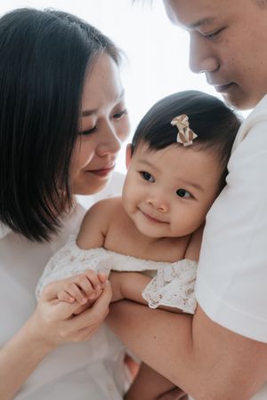 Asian parents lovingly holding their smiling baby girl with a bow in her hair, dressed in white lace.