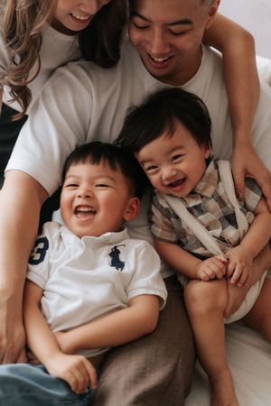 Happy Asian family with two laughing toddler boys sitting together, parents smiling warmly in a bright, cozy setting.