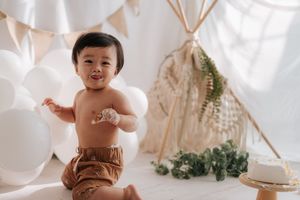 A happy baby in brown bloomers sits among white balloons near a boho teepee backdrop at a first birthday cake smash.