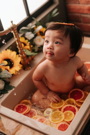 Cute Asian baby sitting in a kitchen sink bath filled with citrus fruit slices and sunflowers nearby.